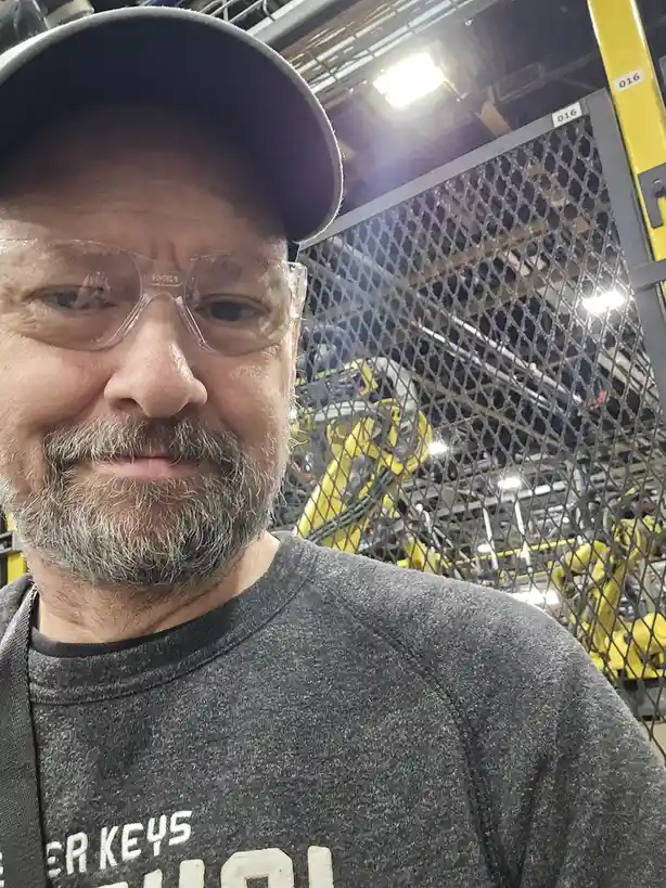 A man wearing a black cap and clear safety glasses smiles at the camera while standing in front of a chain-link fence in…
