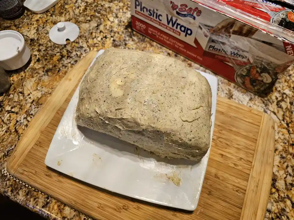 A large, rectangular block of butter, speckled with black pepper, sits on a white plate atop a wooden cutting board on a…
