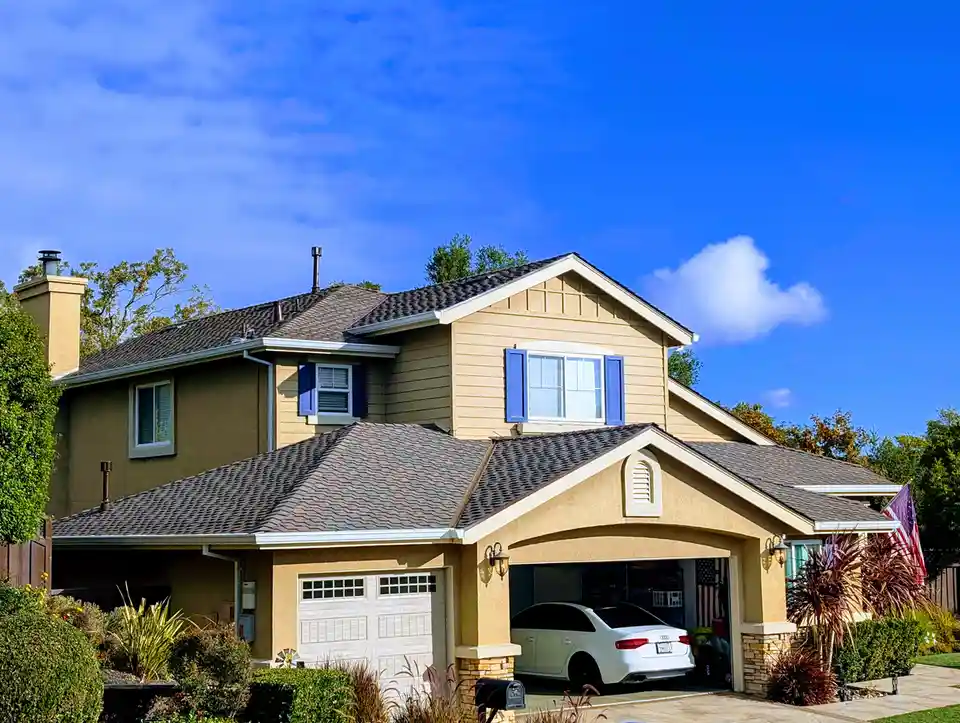 A well-maintained two-story suburban house with beige siding, blue shutters, and a white Audi parked in the open garage. The…