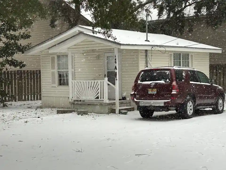 A beige single-story house with a white porch and a maroon SUV parked in front, both covered in snow, with snow visibly…
