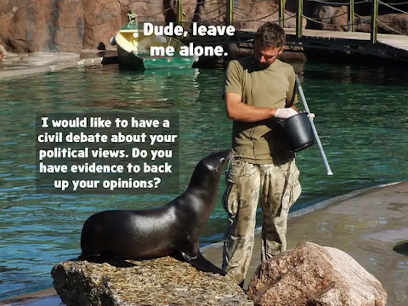 A zookeeper in a green shirt and camouflage pants stands beside a sea lion on a rock near water, holding a bucket and pole…