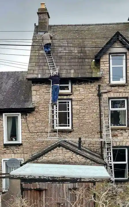 Two men are on ladders attached to a stone house, one near the roof and one lower down, with a third ladder leaning against…
