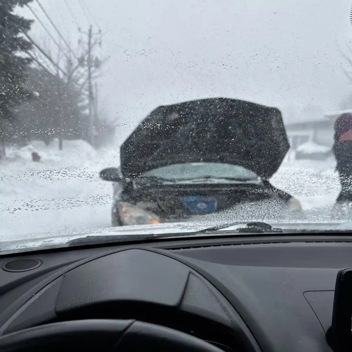 A view from inside a car during a heavy snowstorm, looking out through a rain-splattered windshield at another car with its…