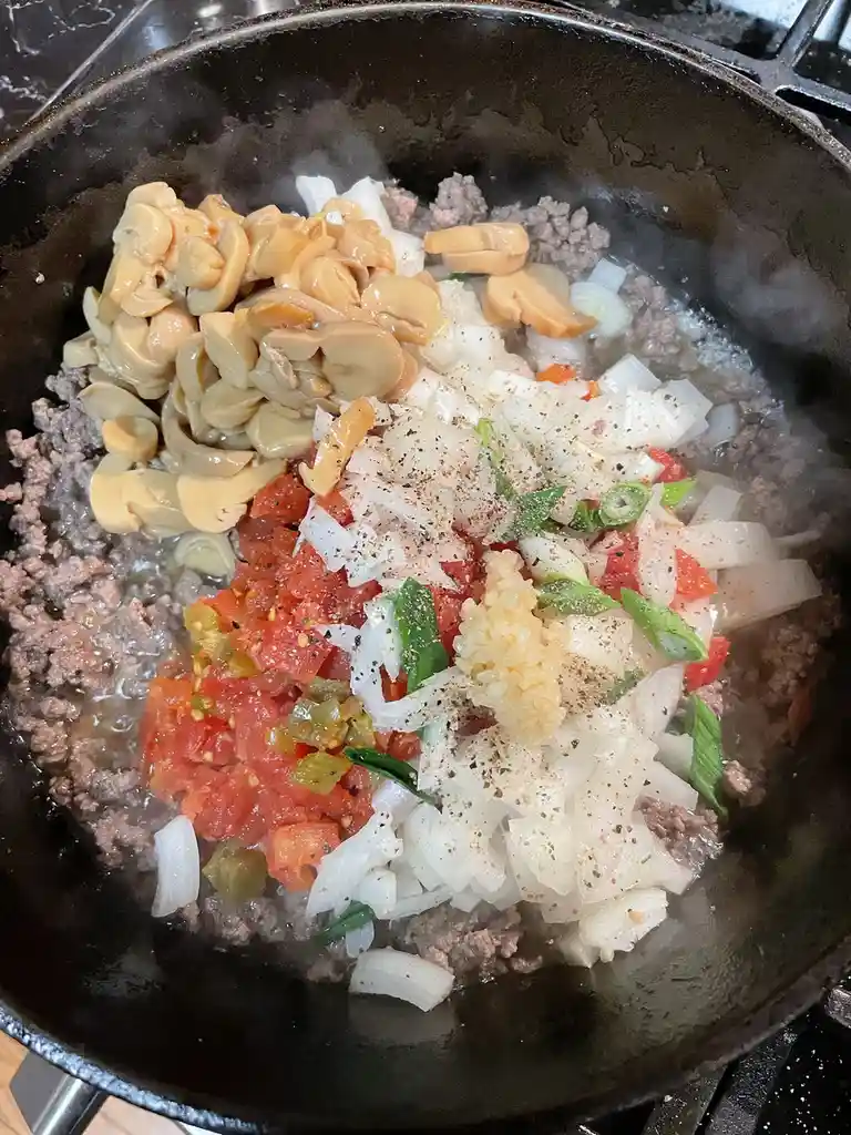 A close-up photo shows a black skillet on a stovetop filled with raw ingredients for a meal, including ground meat, sliced…