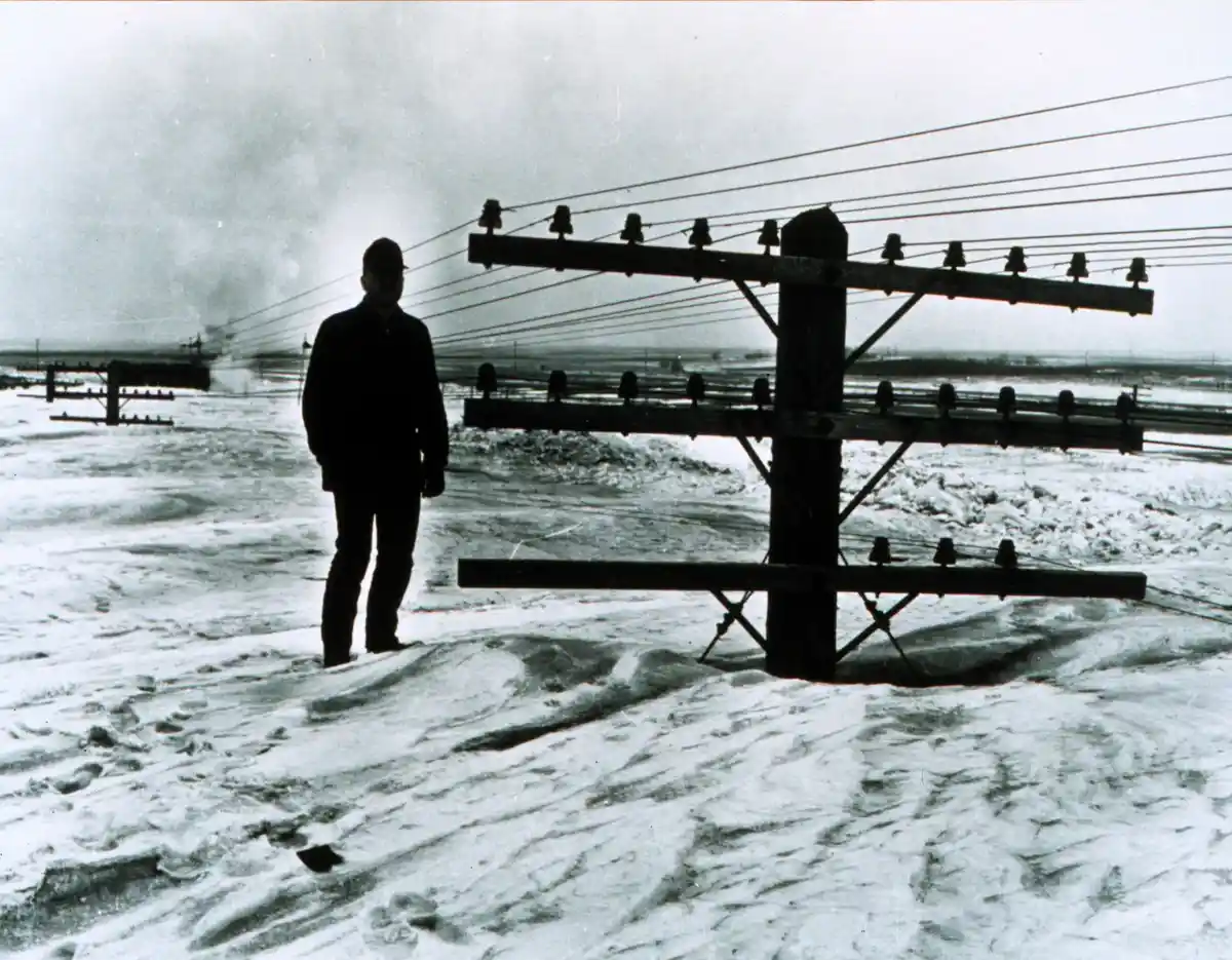 A black-and-white photo shows a silhouetted man standing in deep snow next to a wooden utility pole with wires. The…