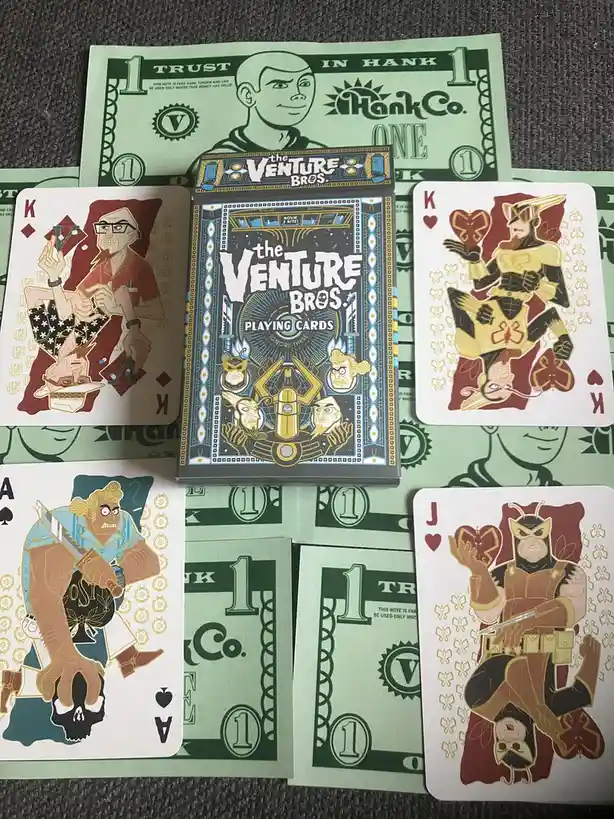 A collection of 'The Venture Bros.' themed playing cards is displayed on top of green 'HankCo.' fake money. The central card…