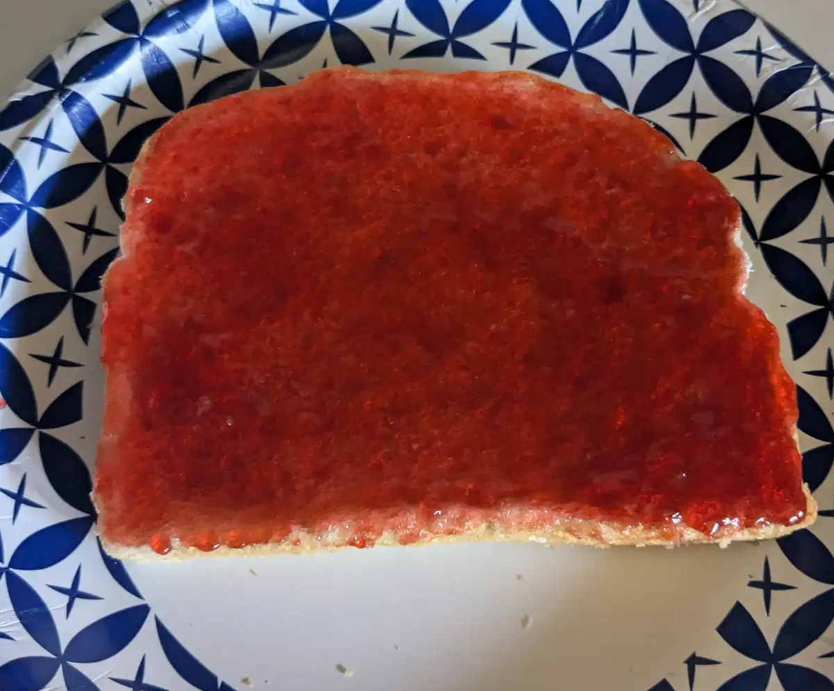 A single slice of white bread, heavily coated with red strawberry jam, sits on a white plate with a blue geometric star…