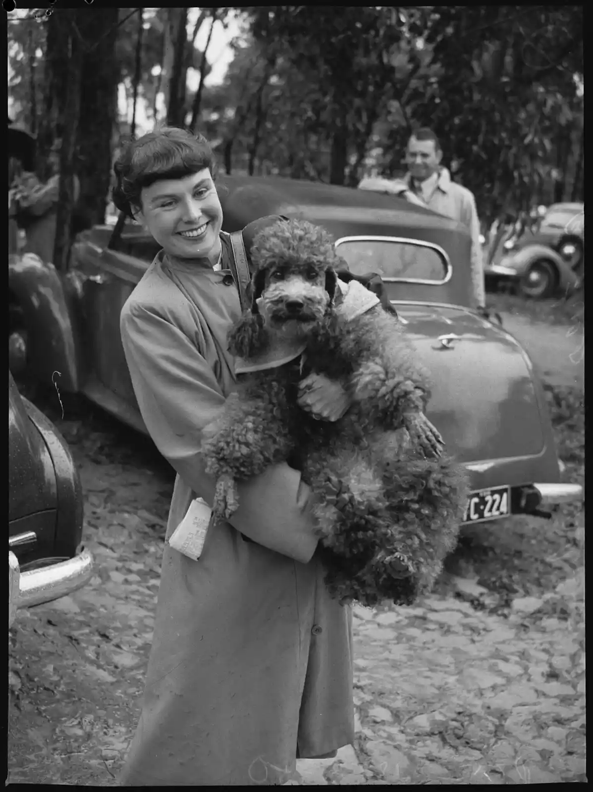 A vintage black-and-white photo shows a smiling woman in a coat holding a large, curly-haired poodle wearing a small jacket…