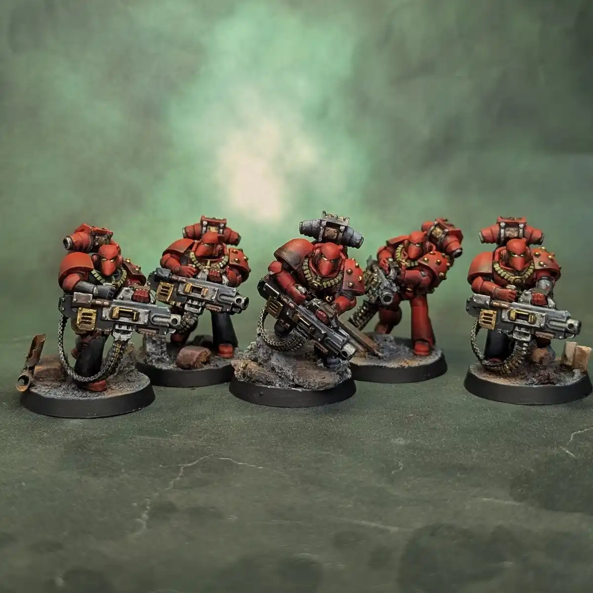 Five painted miniature figures of red-armored, heavily armed space marines stand in a row on rocky bases against a greenish…