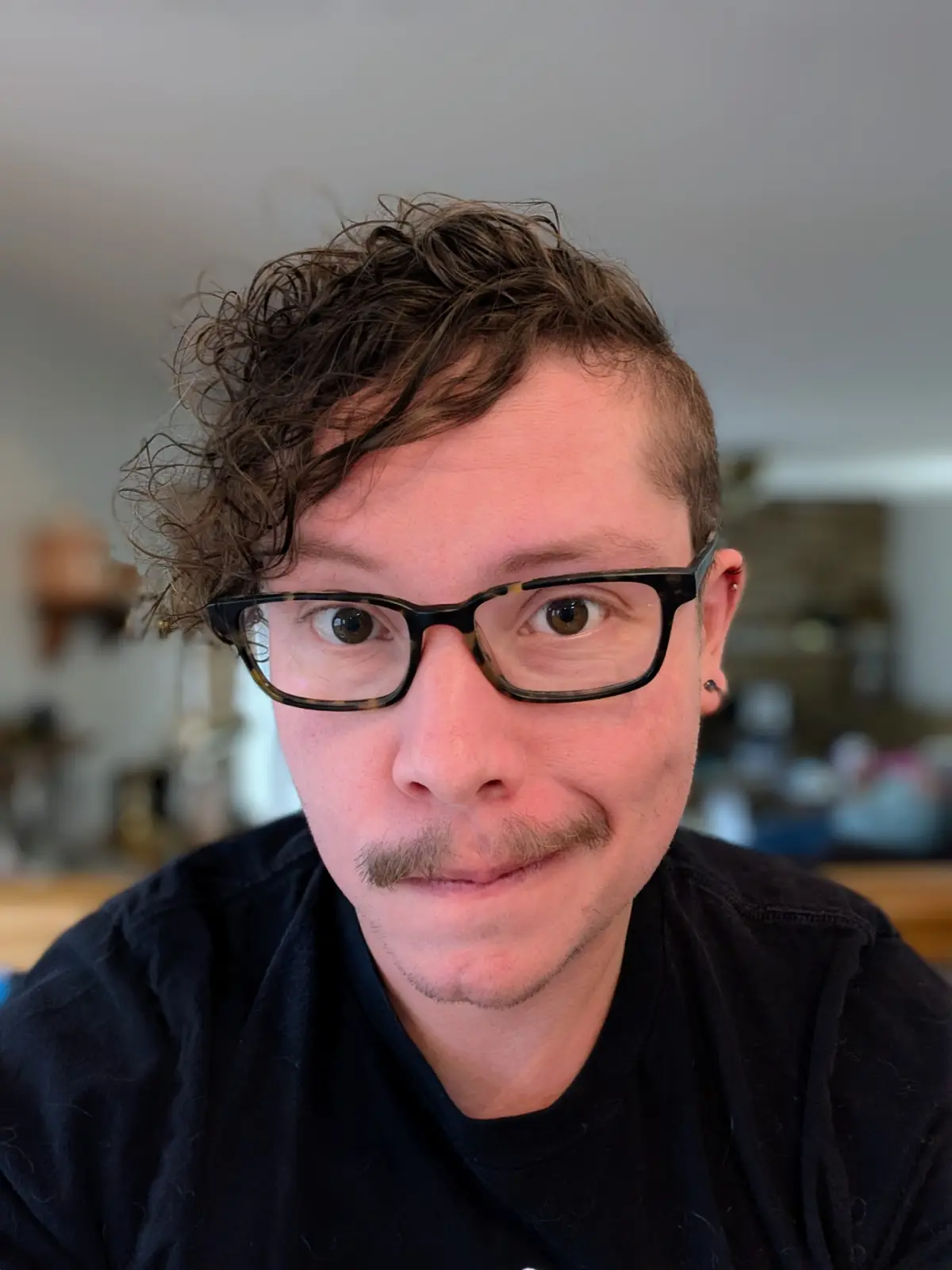 A close-up selfie of a man with curly brown hair, tortoiseshell glasses, and a mustache, wearing a black shirt. He has a…