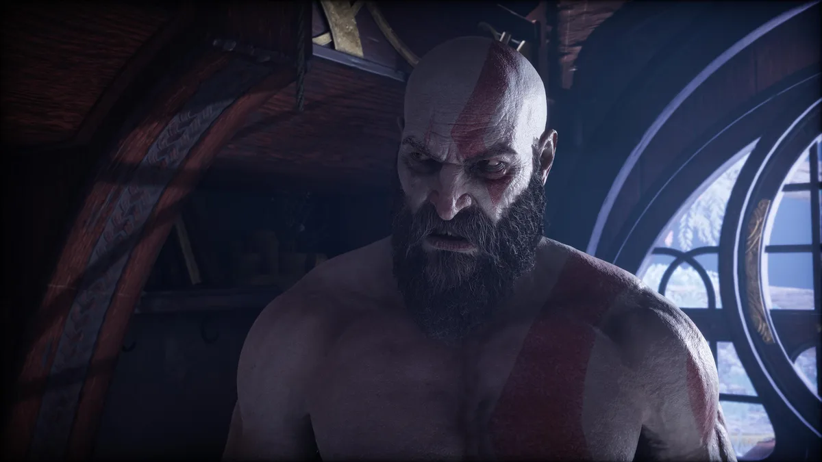 A close-up of the video game character Kratos from God of War, shirtless and looking intensely angry with a furrowed brow…