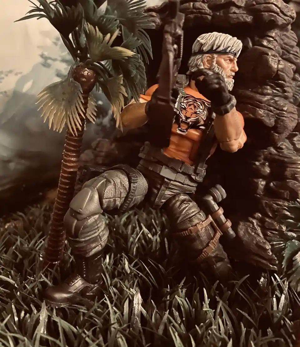 A toy action figure of Big Boss from Metal Gear Solid, with gray hair and beard, wearing an orange tank top with a tiger…