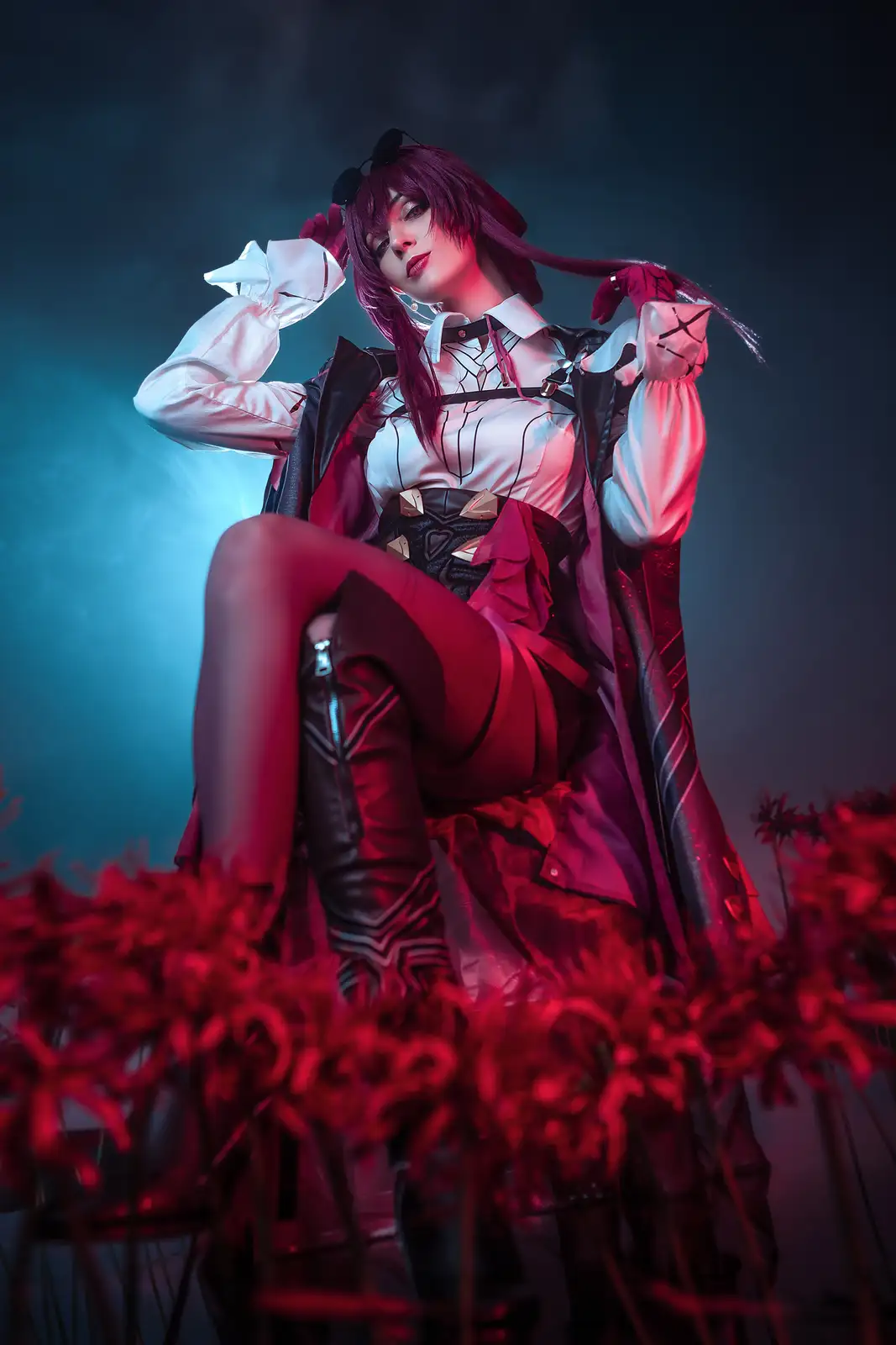 A cosplayer with purple hair and sunglasses perched on her head poses confidently in a white shirt, black vest, and red…