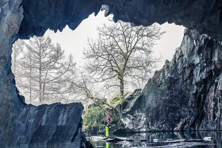 A person wearing a pink hat and bright green pants stands in shallow water inside a large, dark rock cave, looking out at…
