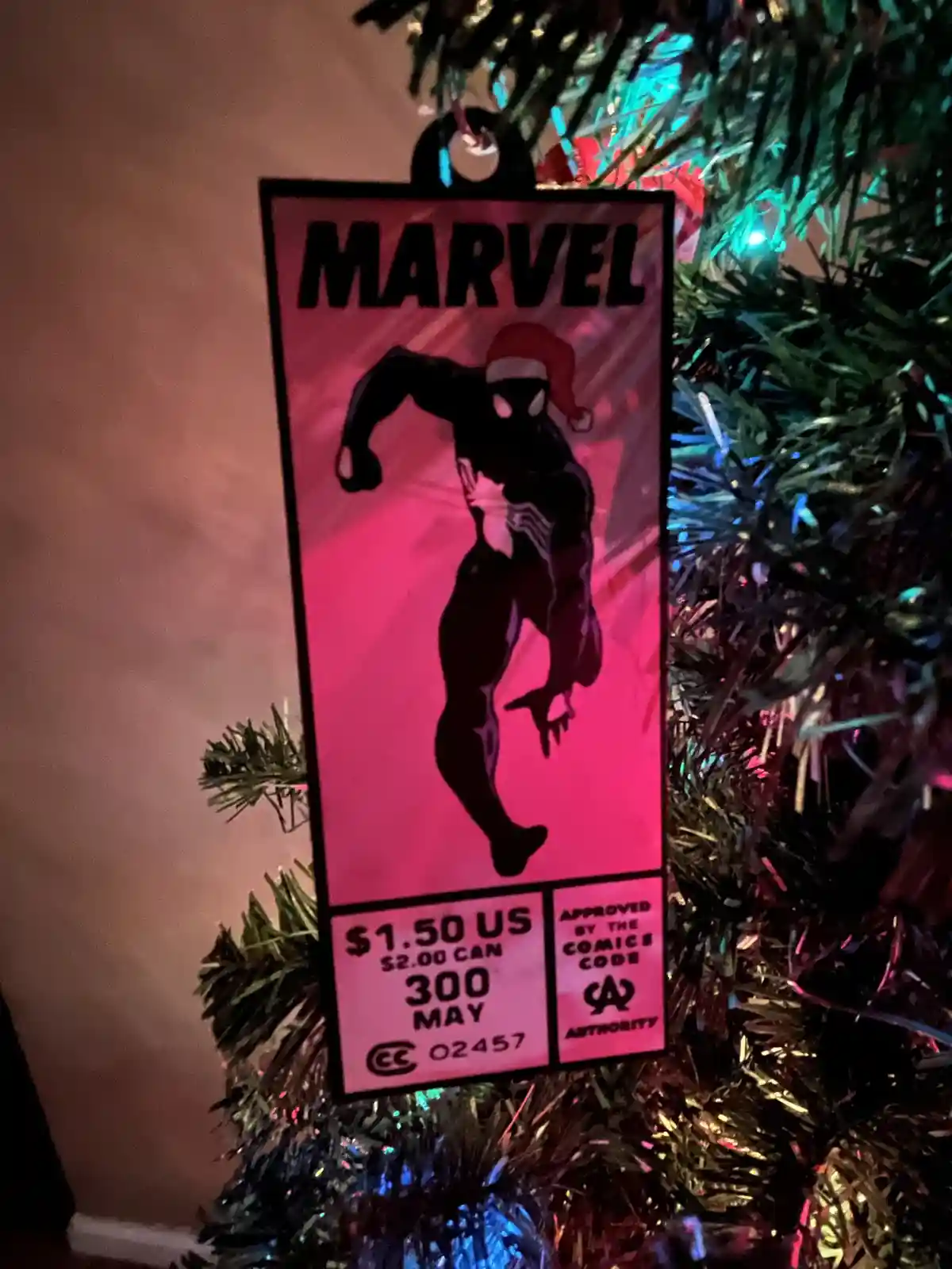 A Christmas tree ornament shaped like a vintage Marvel comic book cover, featuring a black silhouette of Venom wearing a red…