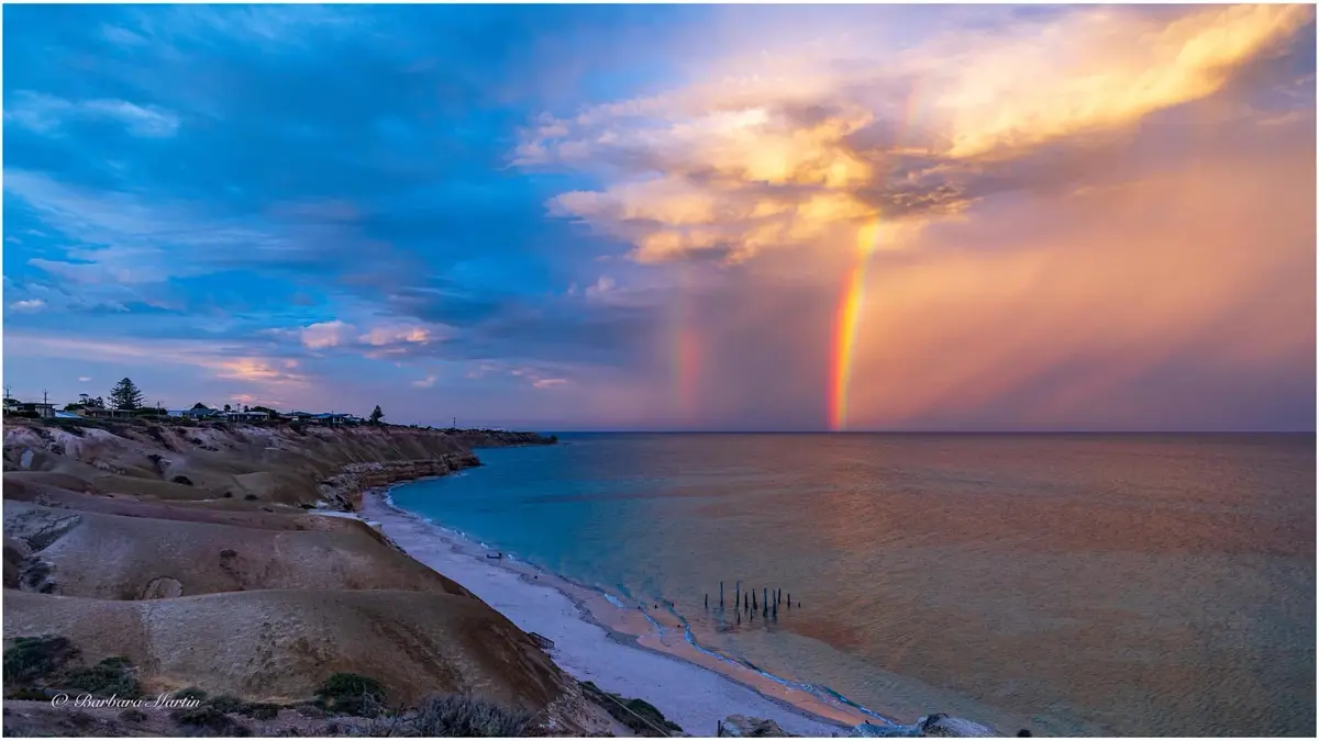 A scenic coastal photograph shows a vibrant double rainbow arcing over the ocean at sunset, with a beach, cliffs, and houses…