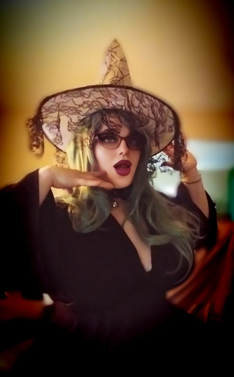 A person with blonde and blue-green hair wears a black lace witch hat and glasses, posing with one hand near their face…
