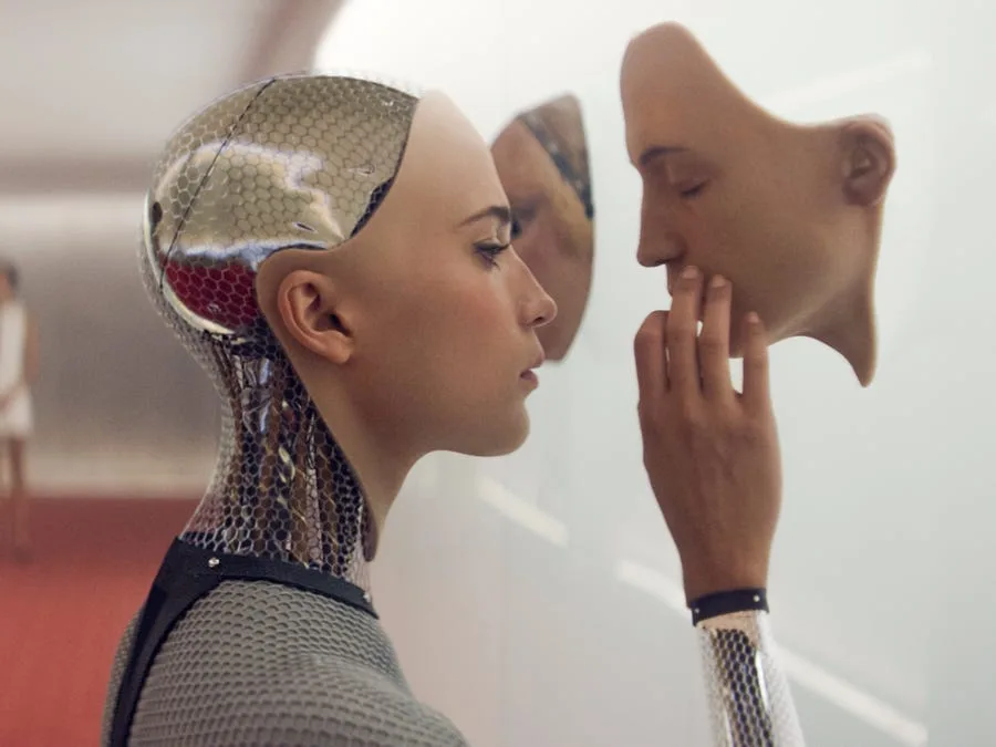 A humanoid robot with a metallic, hexagonal-patterned head and neck, and a human-like face, gazes at its own reflection in a…