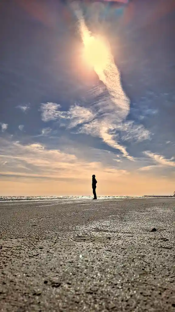 A lone person stands in silhouette on a wet beach at sunset, facing the ocean under a dramatic sky with a bright sun and…