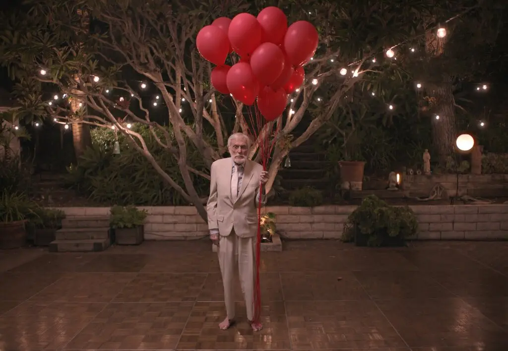 An older man in a light-colored suit stands barefoot on a tiled patio at night, holding a large bunch of red balloons…