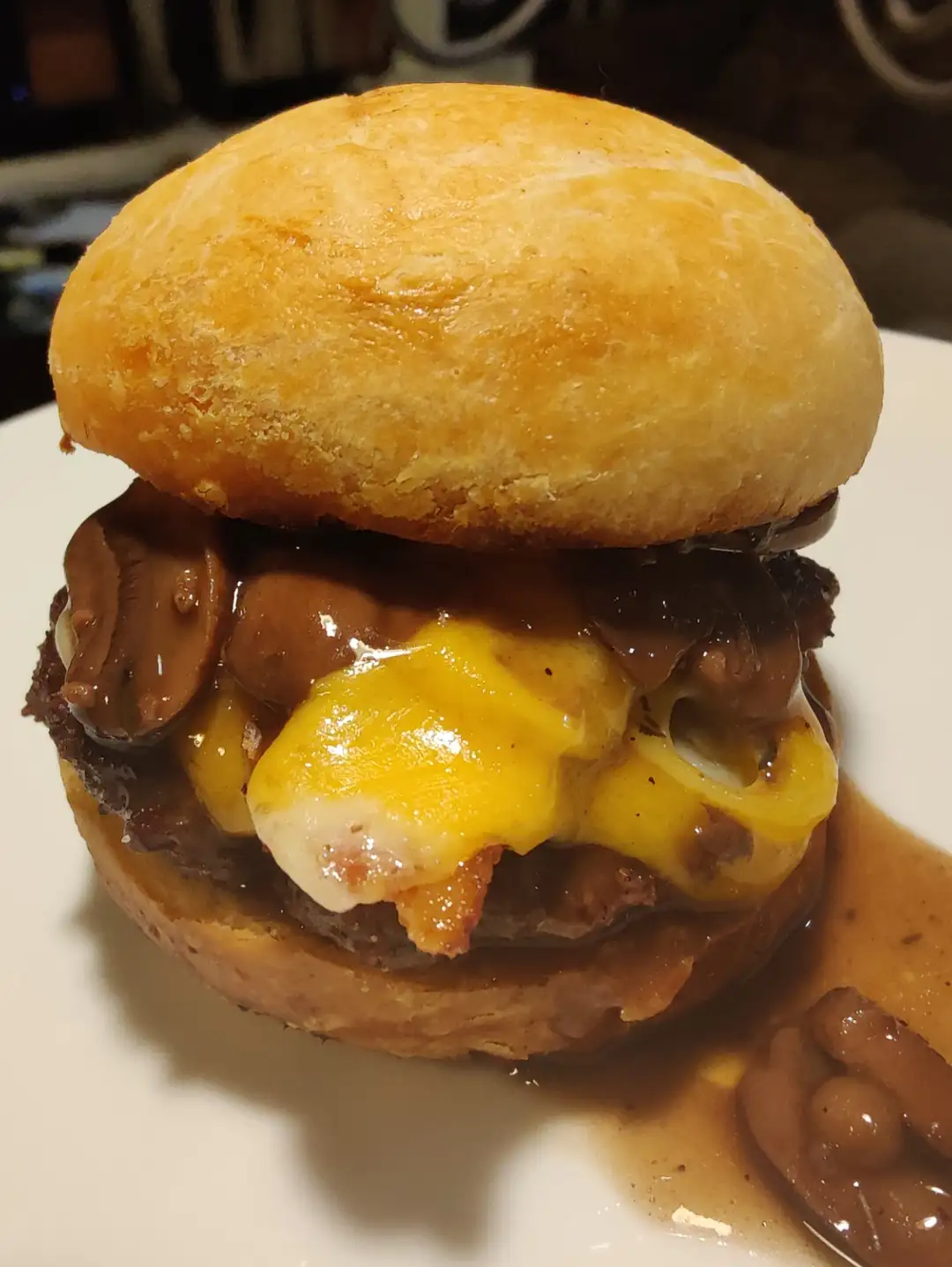 A close-up photo of a burger-style sandwich on a white plate, featuring a golden-brown bun, a beef patty, melted yellow…