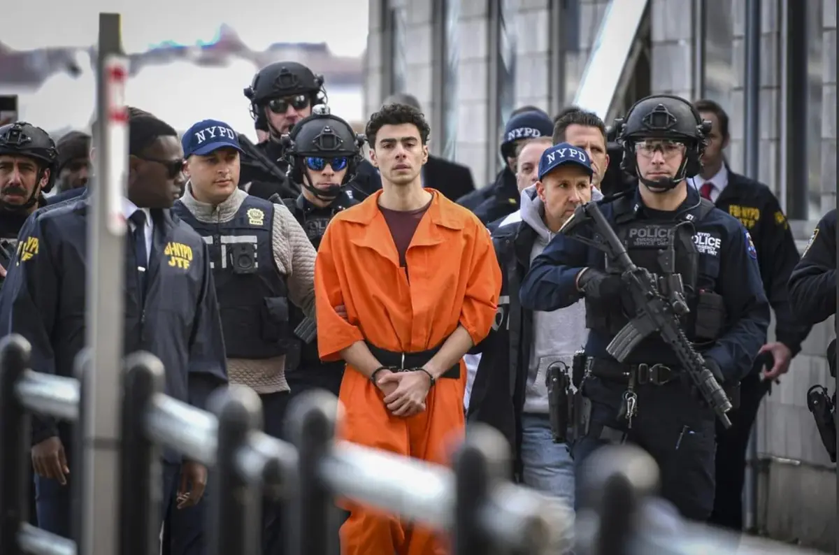 A man in an orange prison jumpsuit stands handcuffed, surrounded by heavily armed NYPD and FBI agents in tactical gear. The…