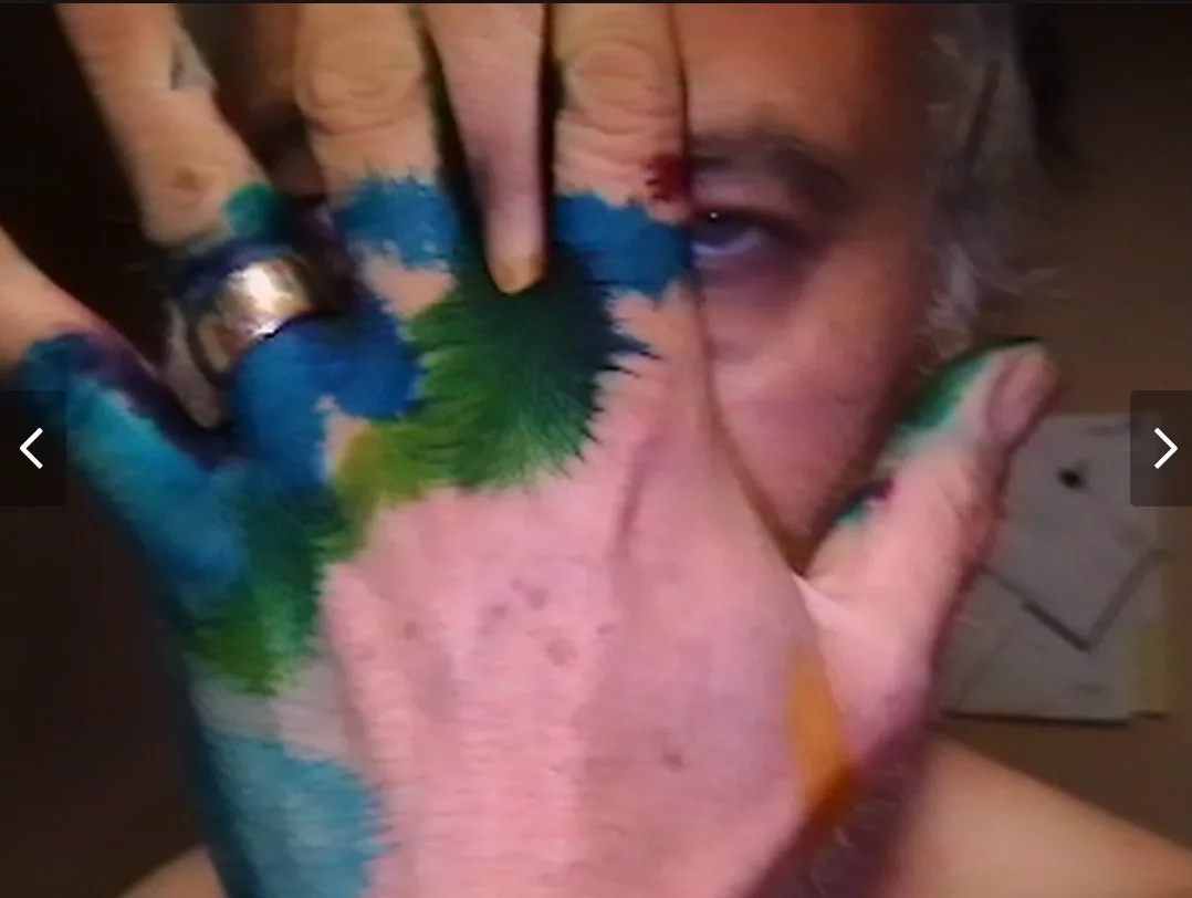 A close-up image shows a person with paint-covered hands partially obscuring their face, revealing only one eye. The hand…