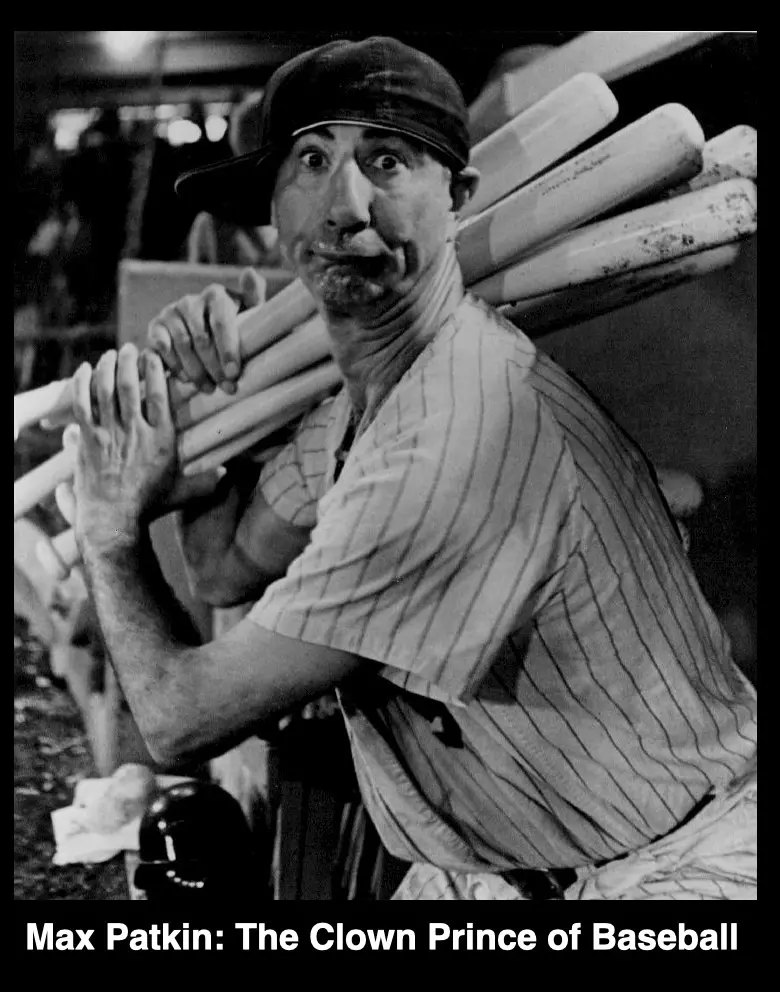 A black-and-white photo shows a man in a pinstripe baseball uniform and cap, carrying a bundle of baseball bats over his…