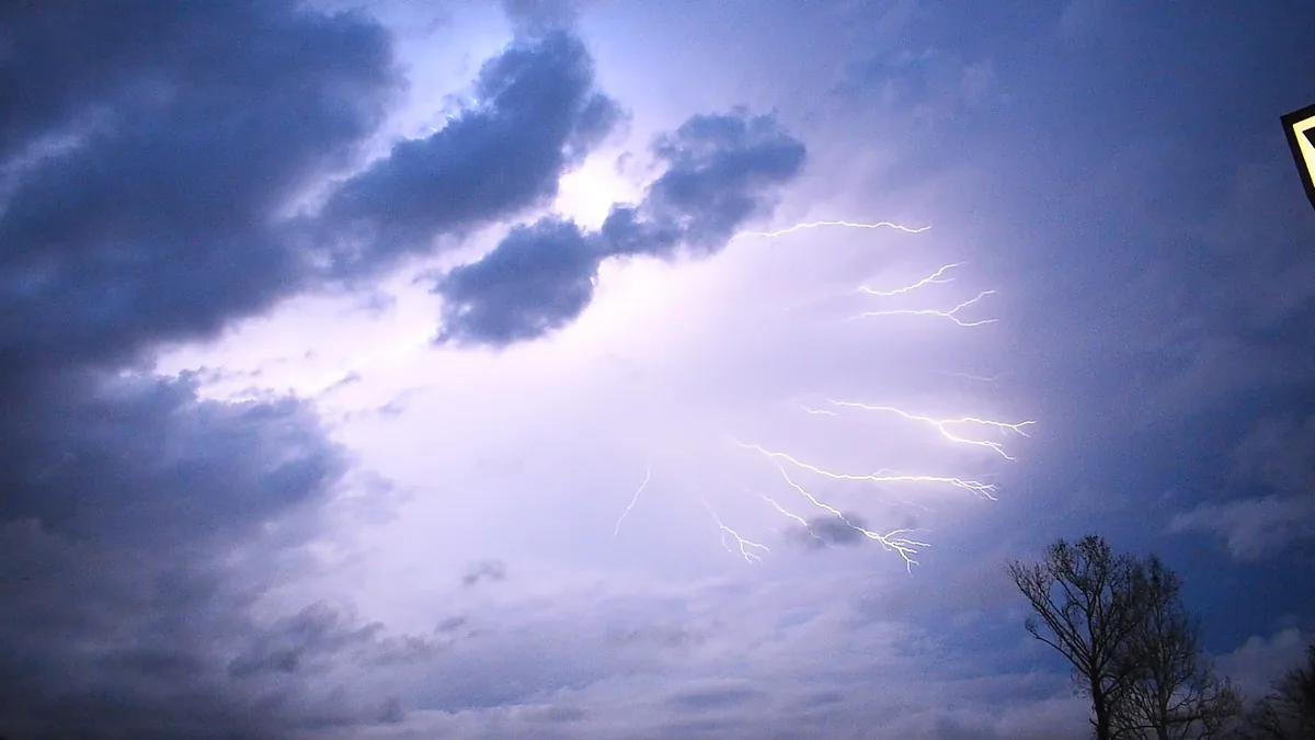 A dramatic sky with dark clouds and bright, branching lightning bolts illuminating the scene. A bare tree silhouette is…