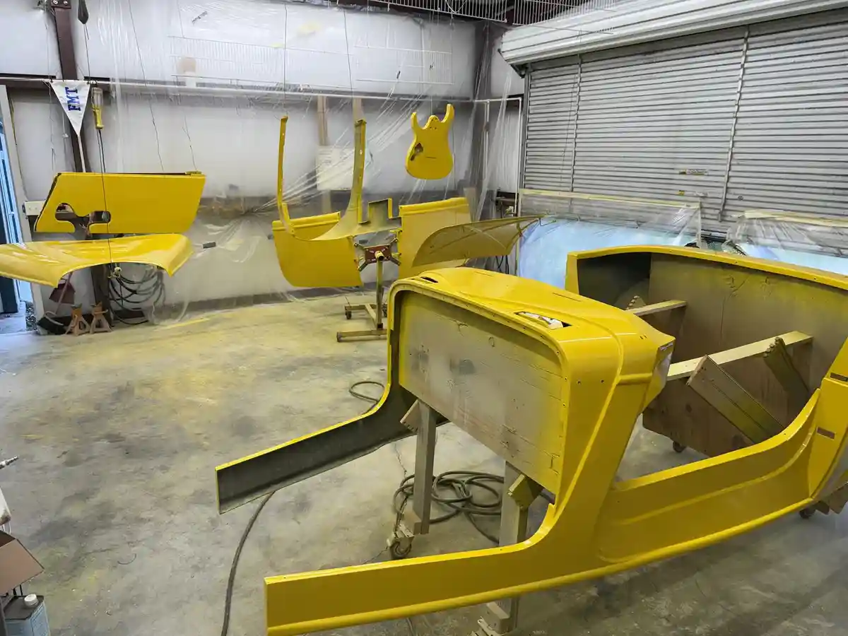 A garage filled with bright yellow car body panels and frames, suspended and resting on stands, with a yellow electric…