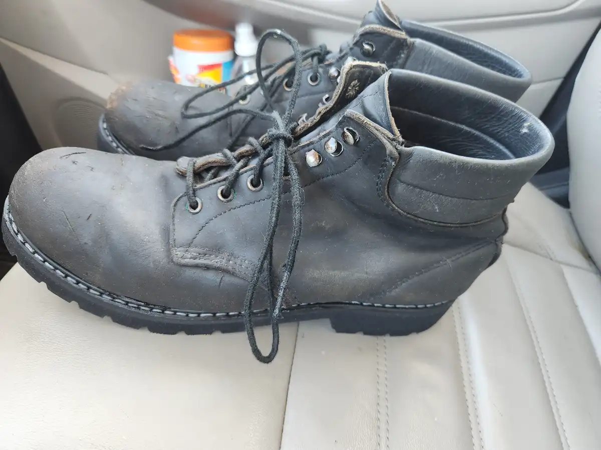 A pair of worn, dirty black work boots with untied laces sits on a light-colored car seat. In the background, a small…