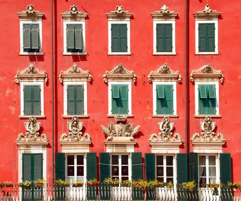 A symmetrical facade of a red building with multiple windows, each framed in white and topped with ornate carvings. All…