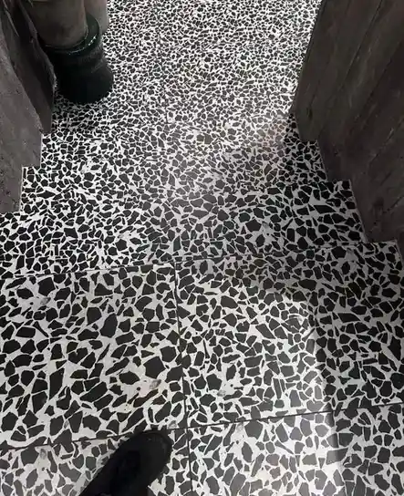 A close-up photo of a black-and-white terrazzo floor with visible grout lines, showing a person's black shoe at the bottom…