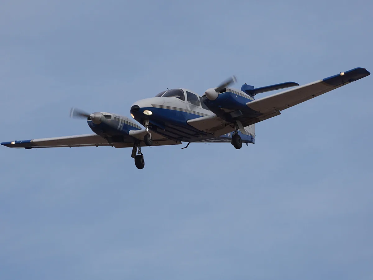 A blue and white twin-engine propeller airplane is flying low against a clear blue sky, with its landing gear down and…