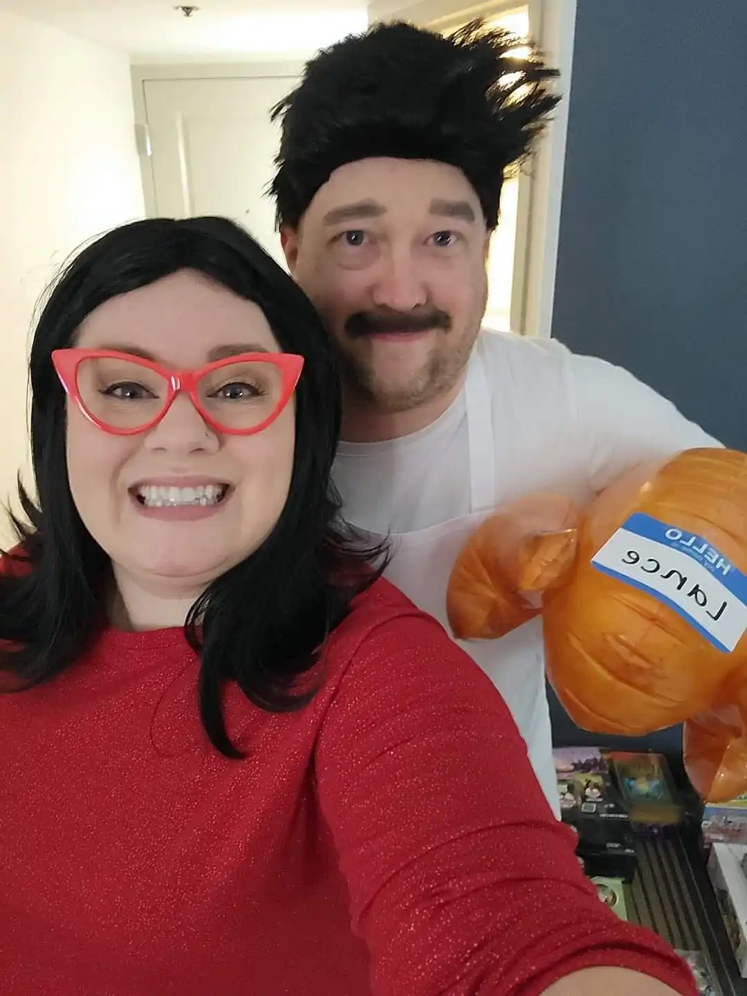 A man and woman pose for a selfie, the man wearing a spiky black wig and fake mustache while holding an orange inflatable…