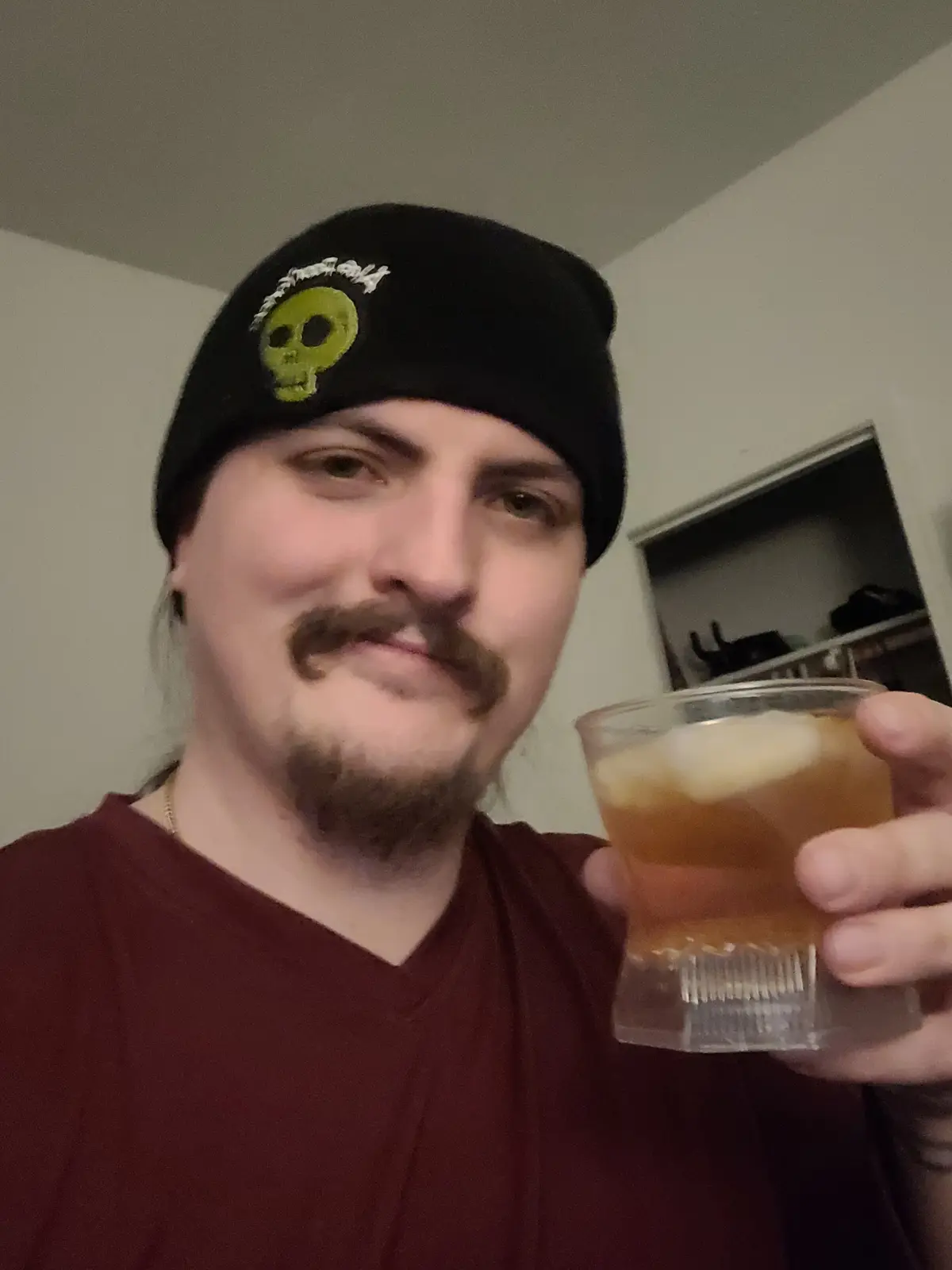A man with a mustache and goatee wearing a black beanie with a green skull logo holds up a plastic cup with a drink and ice…