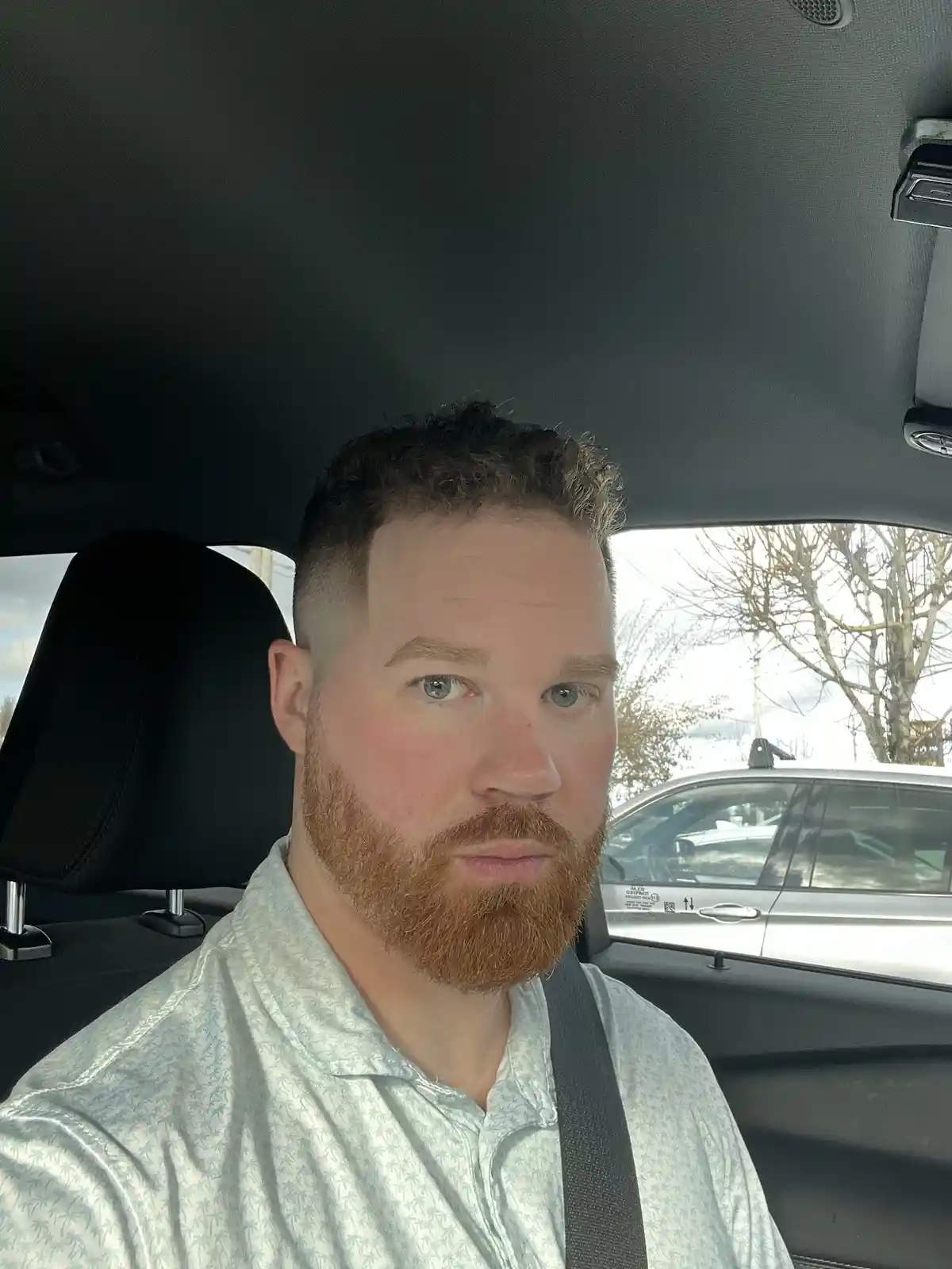 A man with a reddish beard and blue eyes sits in the driver's seat of a car, wearing a light-colored shirt with a palm tree…