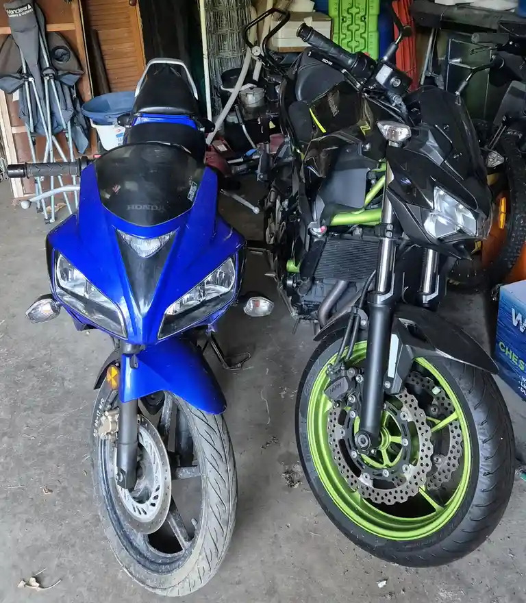 Two motorcycles are parked side-by-side in a cluttered garage. The left one is a bright blue Honda with a standard front…