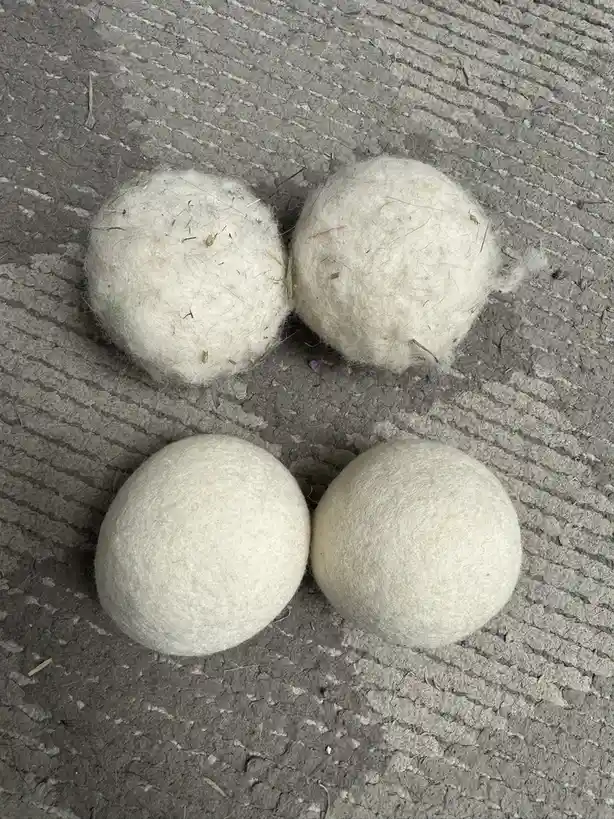 Four off-white wool dryer balls sit on a gray textured carpet. The top two balls are visibly covered in embedded lint and…