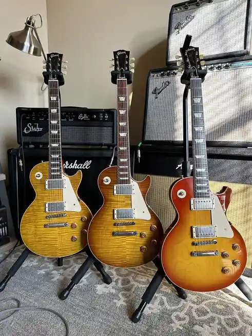 Three Gibson electric guitars with sunburst finishes stand upright on stands in front of several amplifiers, including…