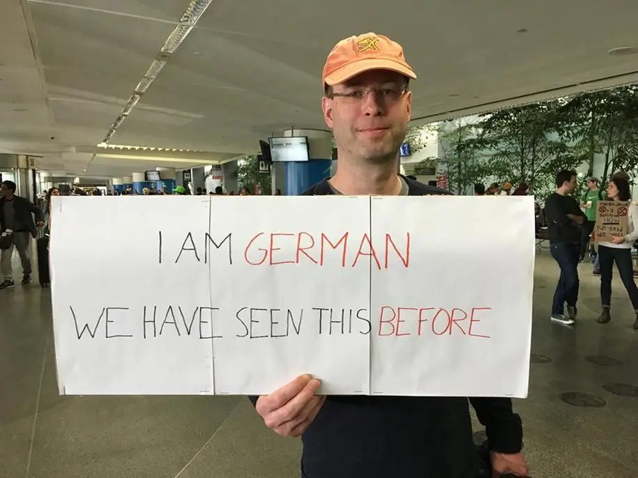 A man wearing an orange cap and glasses holds a large white sign in an airport terminal. The sign reads 'I AM GERMAN' in…