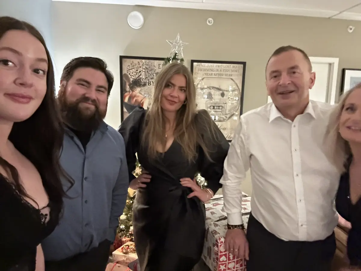 A group of five people pose for a photo in a festively decorated room with a Christmas tree and holiday-themed wall art. The…