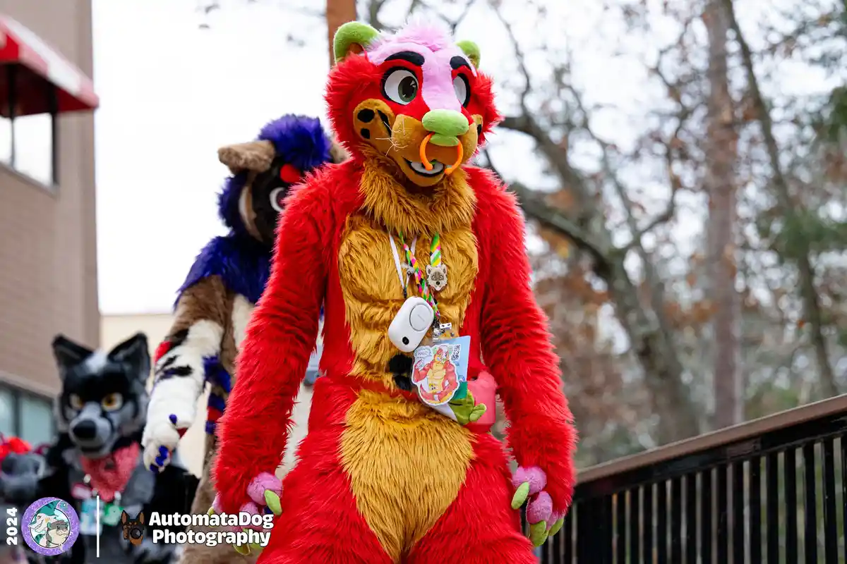 A person in a vibrant red and yellow fursuit resembling a stylized lion or tiger with green ears and nose, wearing a white…