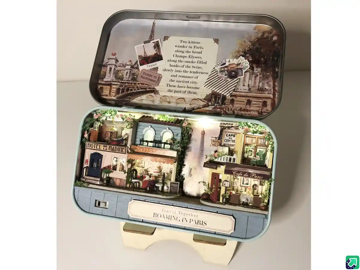 An open, light-blue tin box displays a miniature Parisian street scene with tiny buildings like a hotel, patisserie, and…
