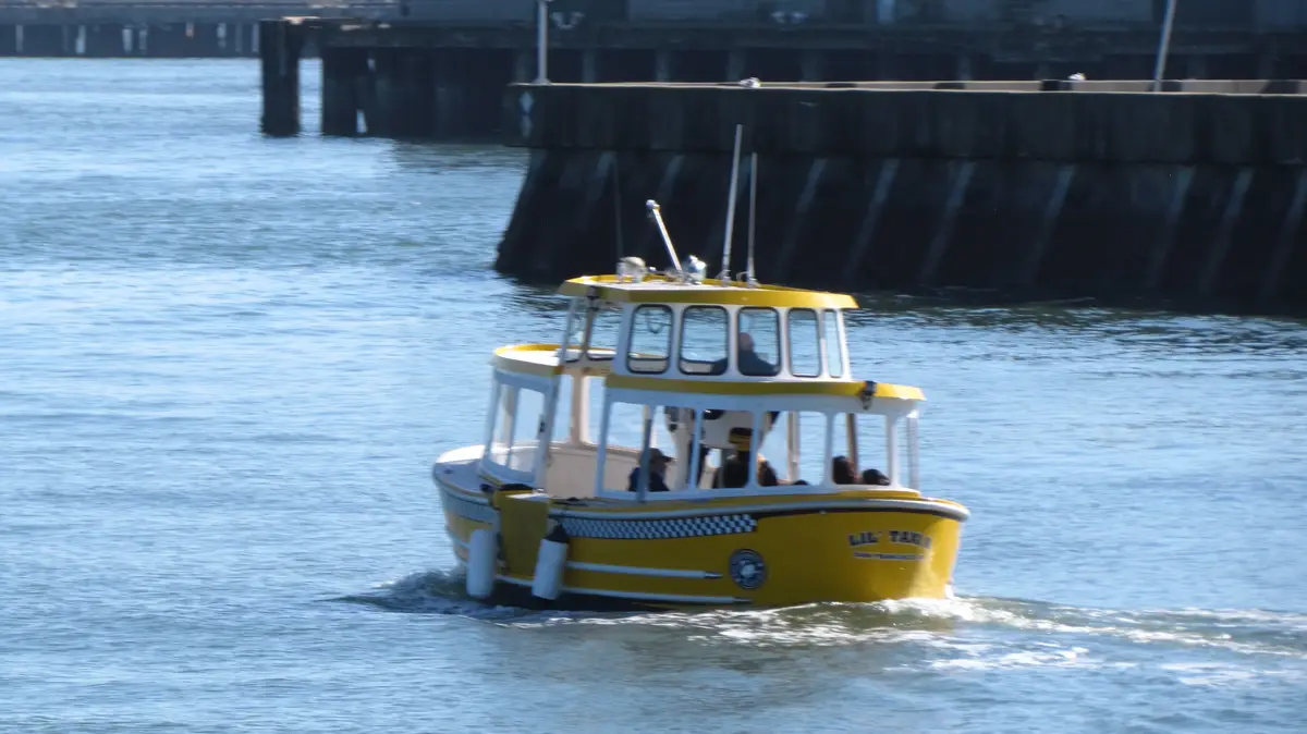A yellow water taxi boat with a checkered stripe and 'LIL' TAXI' written on its bow is moving through blue water near a…