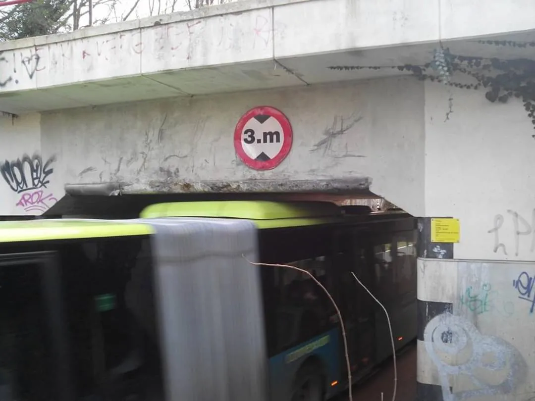 A blurry photo shows a dark green and lime-green bus driving under a concrete overpass. On the wall above the bus, a…