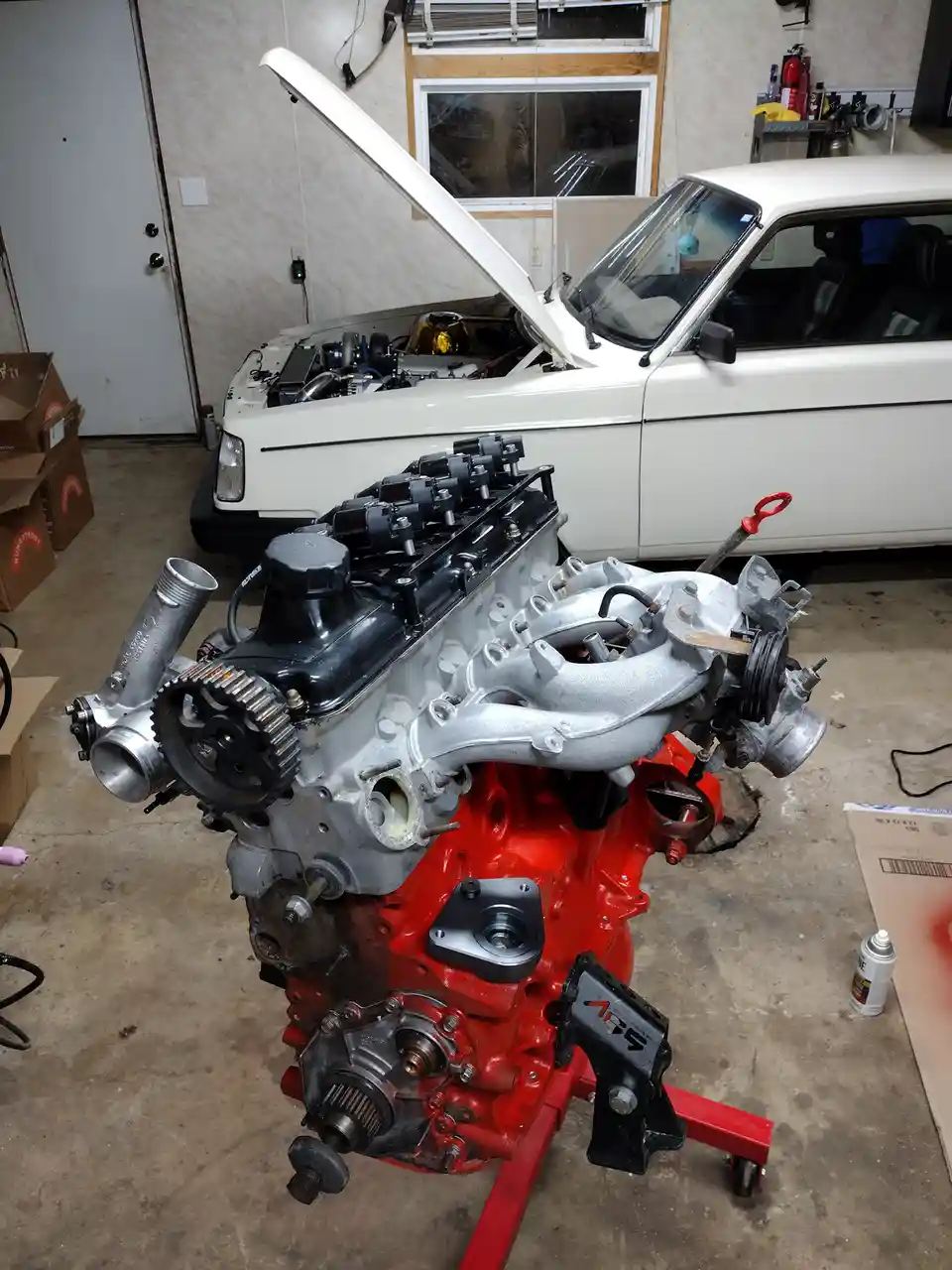 A partially assembled car engine, painted red at the base and silver on top, sits on a red stand in a garage. Behind it, a…
