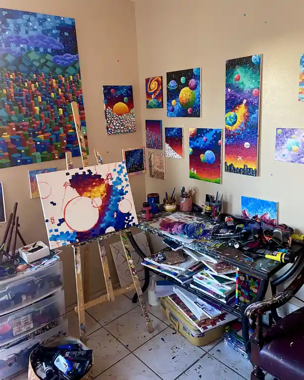A cluttered artist's studio filled with colorful, pixelated paintings on easels and walls, paint tubes, brushes, and…
