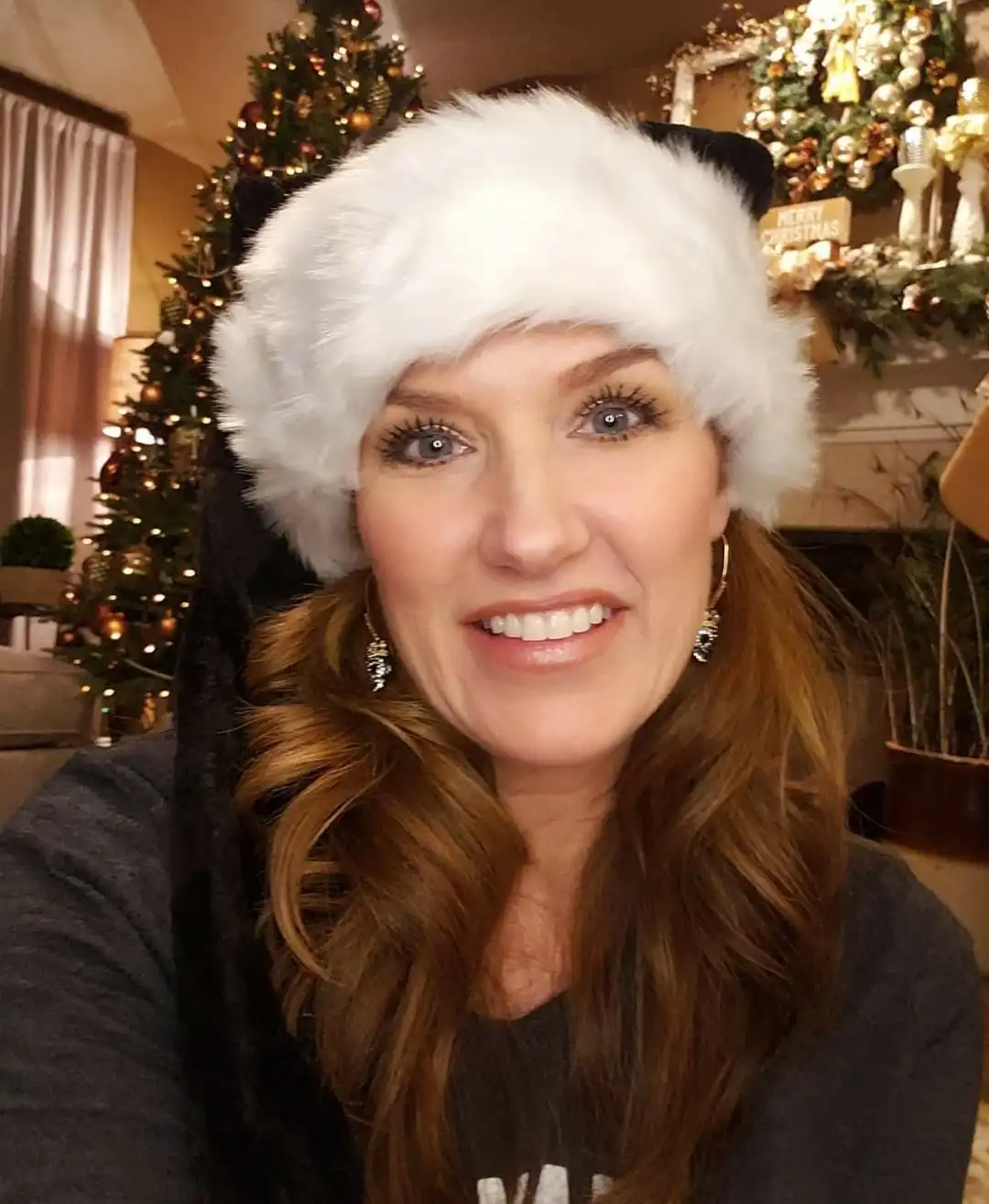 A woman with long brown hair and bright makeup smiles at the camera while wearing a white furry Santa hat with black cat…