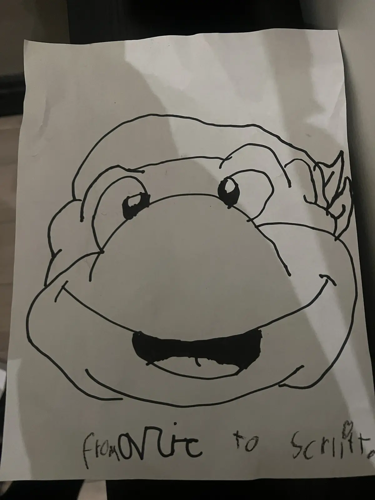 A simple black-and-white line drawing on paper depicts a cartoonish frog-like face with large eyes, a wide smile, and a…